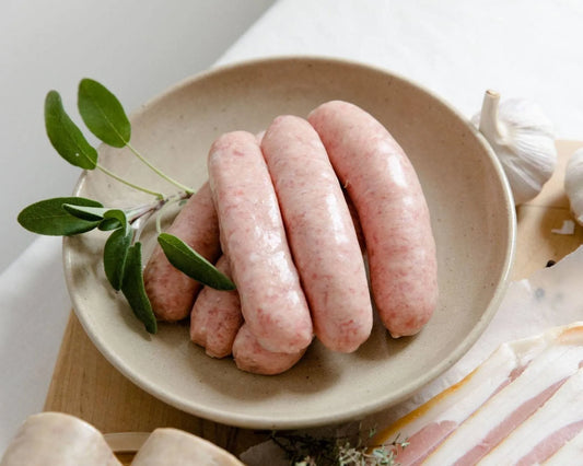 Primrose Herd pork sausages, Cumberland style, shown raw on a wooden cutting board with herbs and seasoning.