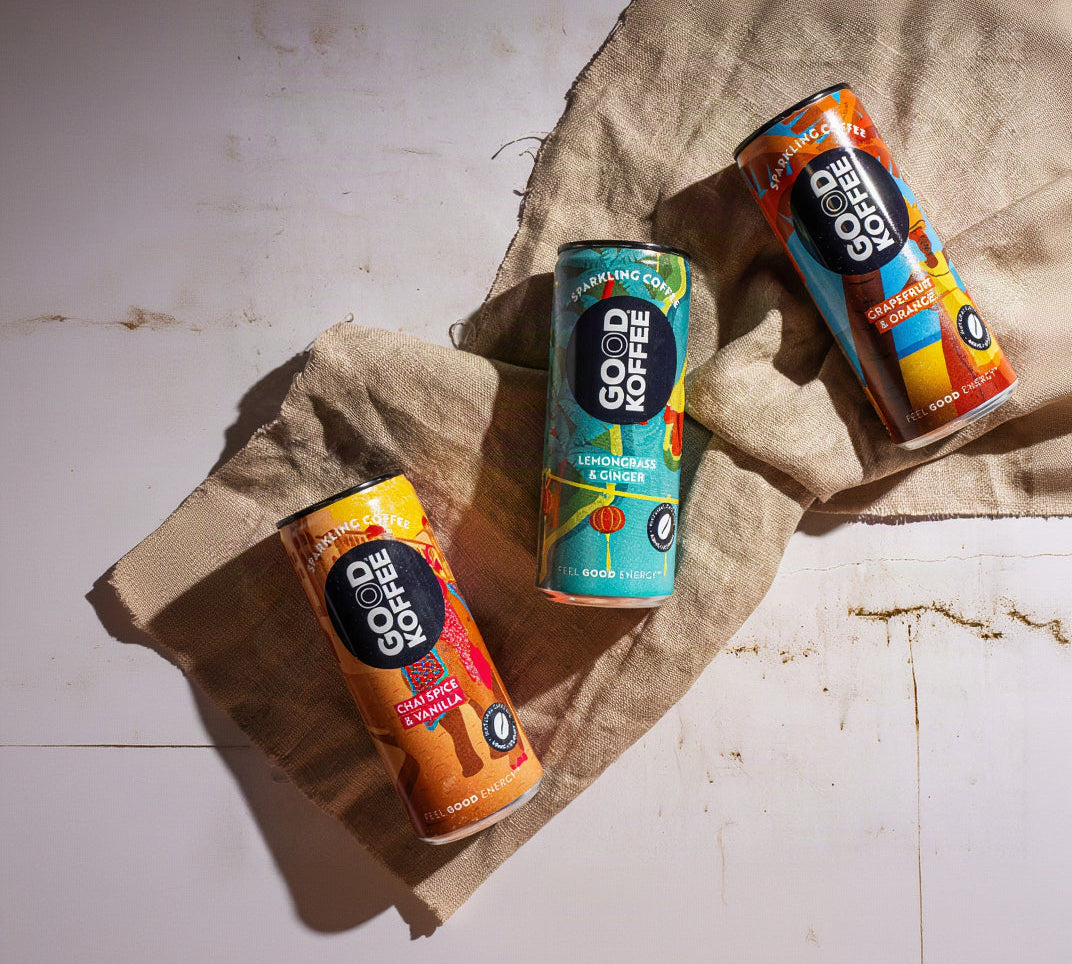 Three cans of GOOD KOFFEE Sparkling Coffee drink arranged on a beige linen cloth over a white surface, with strong directional sunlight casting shadows.