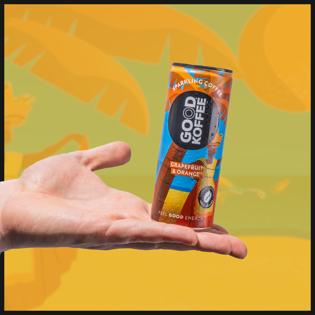 A can of GOOD KOFFEE sparkling coffee with grapefruit and orange flavor, held in a person's hand against a background with palm leaves and yellow and orange colors.