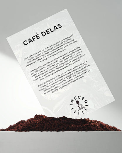 Recent Beans Nose To The Grindstone Medium Roast Coffee in a bag with coffee beans and a steaming cup on a wooden table.