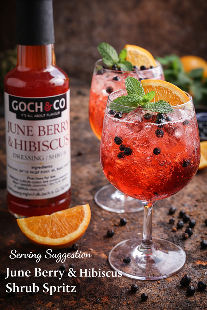 Bottle of Goch & Co June Berry & Hibiscus dressing with two glasses of a red drink garnished with mint and orange slices on a rustic surface.