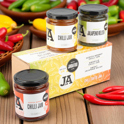 Jars of Janda food Chilli Jams on a wooden surface with a selection of chilli peppers in the background.