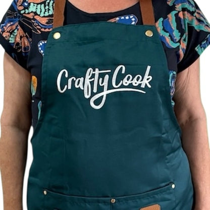 Teal Crafty Cook apron with logo and multiple pockets, designed for cooking and baking, made of durable fabric.