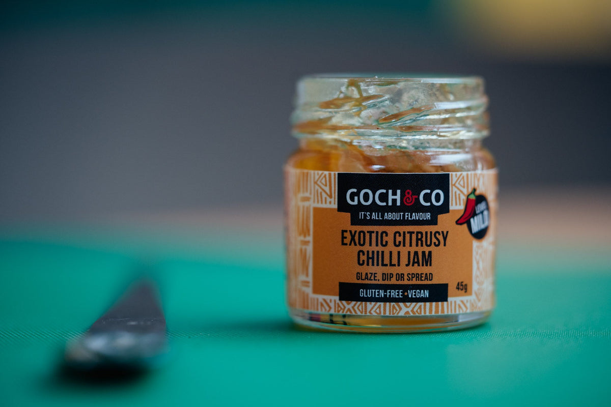 Spicy Goch & Co Exotic Citrusy Chilli Jam jar with vibrant red label and fresh citrus slices on a wooden surface.