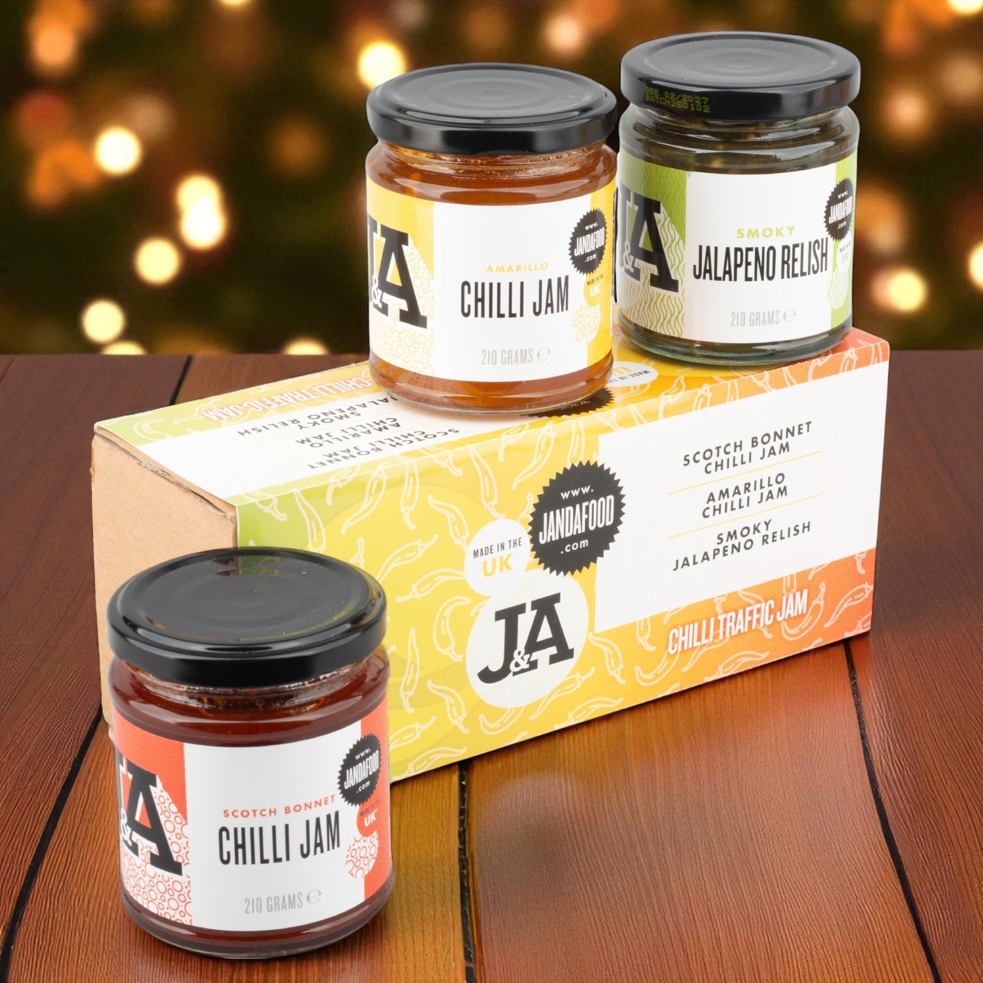Three jars of J&A Chilli Jam and Smoky Jalapeno Relish on a wooden surface with a bokeh background.