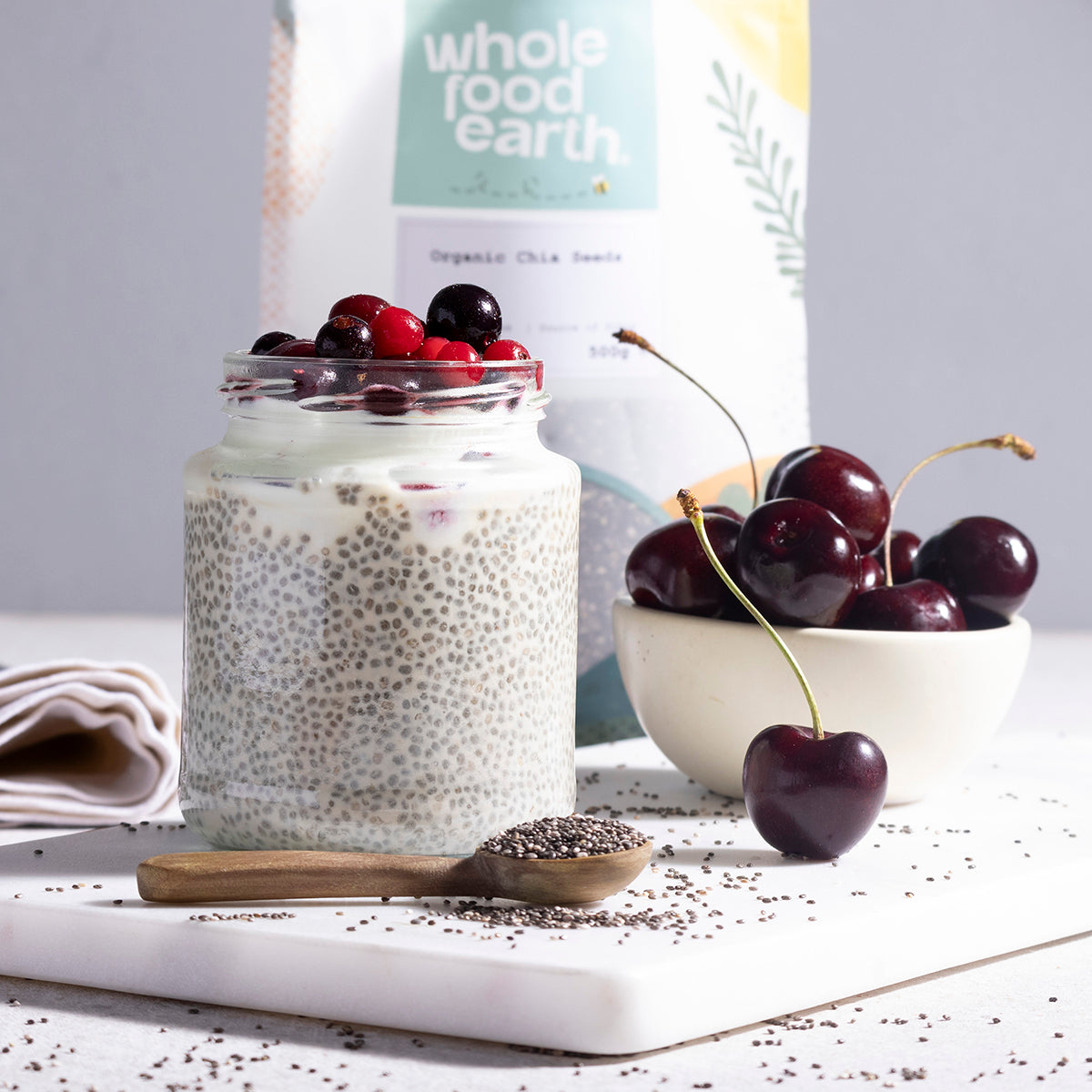 Organic chia seeds from Whole Food Earth displayed in a clear glass jar with a spoon, on a wooden surface with fresh fruit.