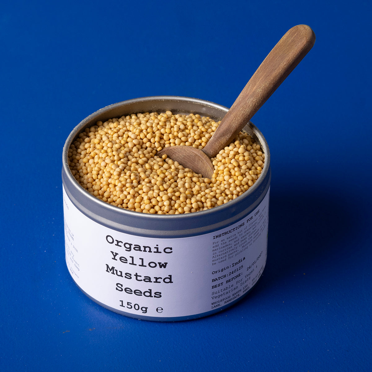 Organic yellow mustard seeds from Wild Elephant, 150g, displayed in a clear glass jar with seeds spilling out, on a wooden surface.