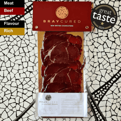 Thin slices of Bray Cured Classic Bresaola, a spicy Italian charcuterie, displayed on a wooden board with herbs and accompaniments.