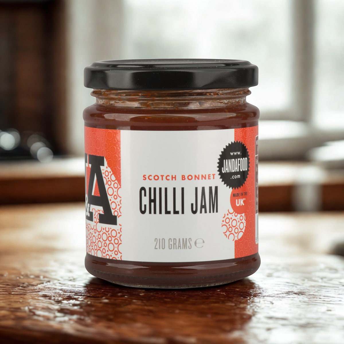Janda Scotch Bonnet Chilli Jam jar showing the label, with a spoon of bright red spicy-sweet chili jam beside it.