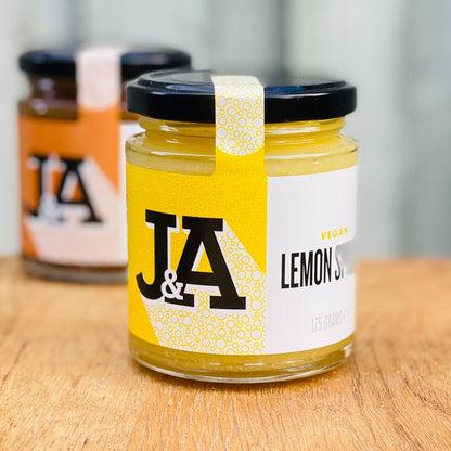 Jar of Janda vegan lemon spread with fresh lemon slices and spread on a wooden table, showcasing its bright yellow color.